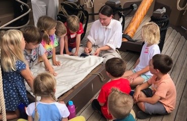 Children watch a costumed interpreter mend a sail