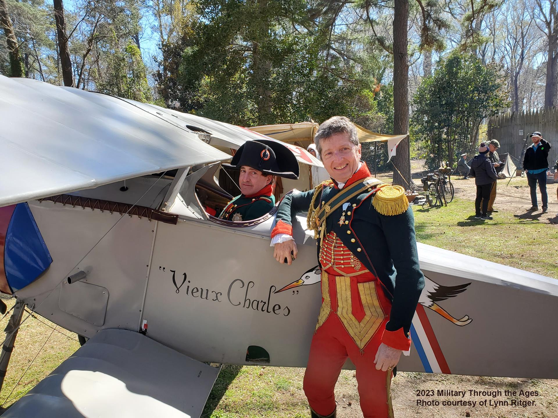 Two men in French dress uniforms are near a replica WWI airplane