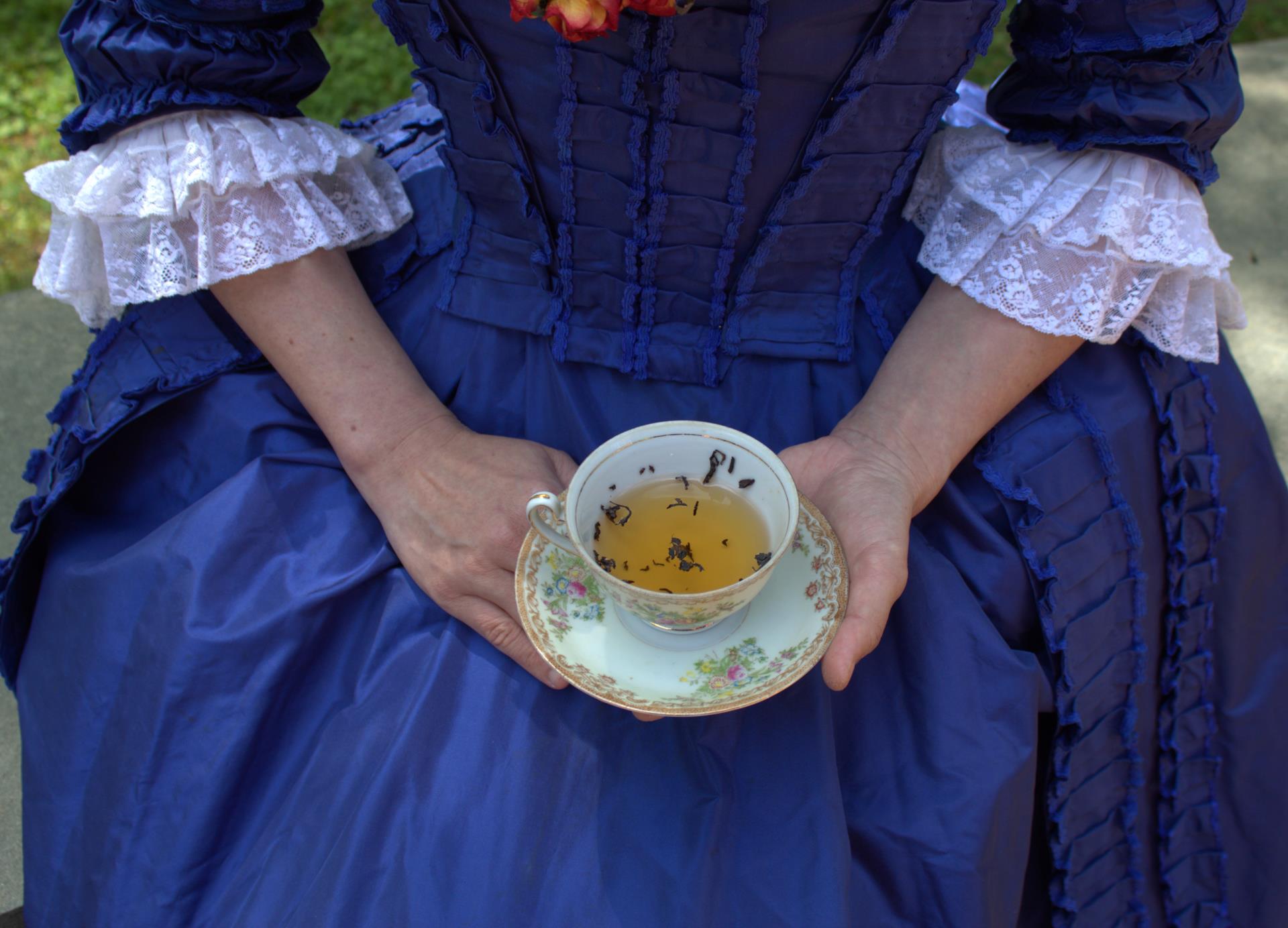 A woman in a Colonial grown holds a teacup and saucer