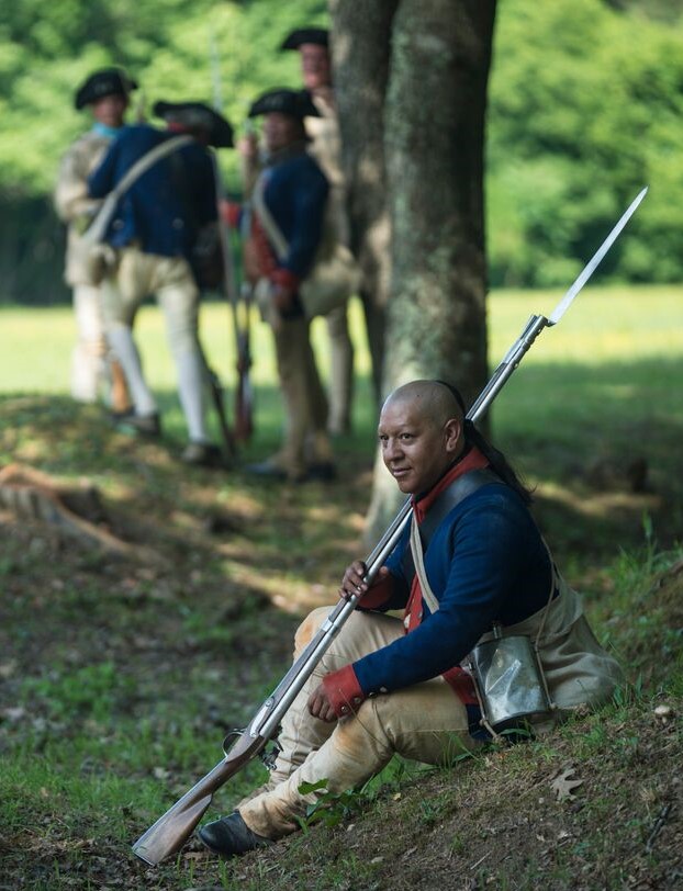 An Indigenous man in a Continental Army uniform sits under a tree